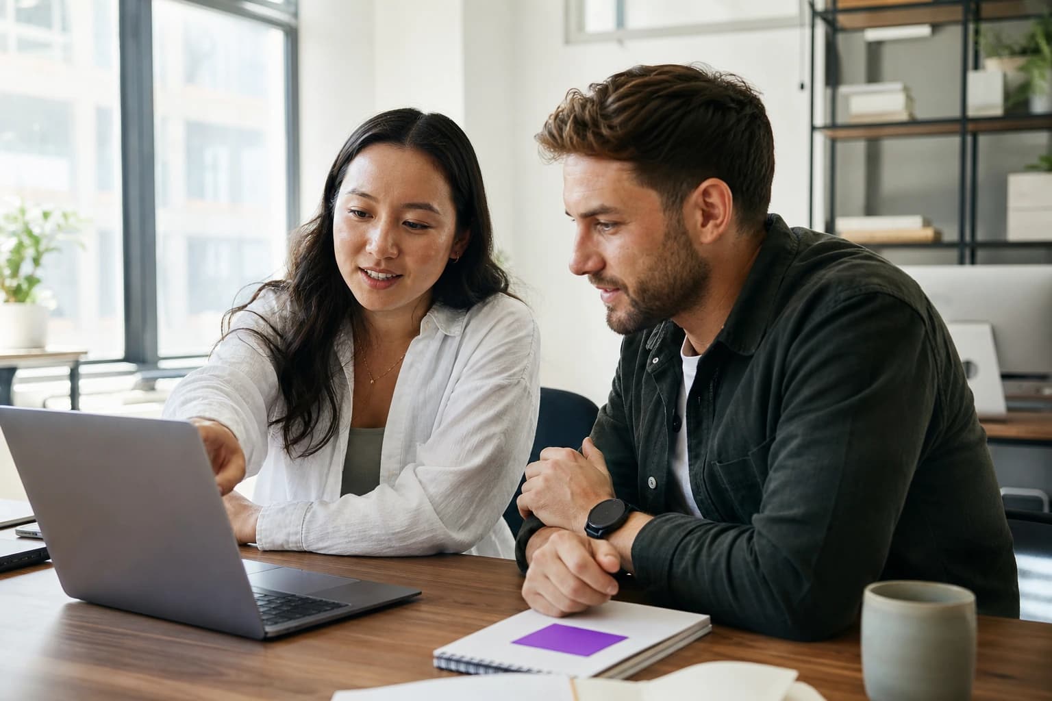 Two colleagues at a bright office desk, one pointing at a laptop screen as they review a draft together before it is approved