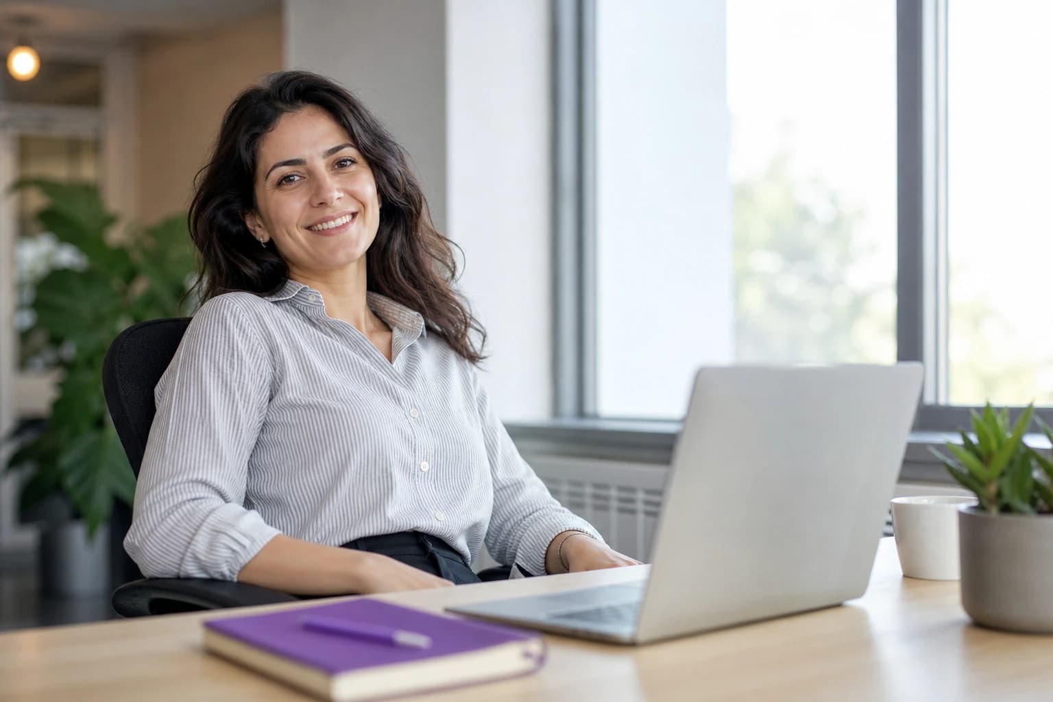 A professional relaxed at her desk beside an open laptop, with a purple notebook and pen on the desk